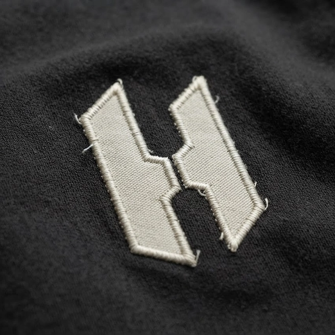 Nærbilde av Håndverker Mobekks H-logo brodert i beige tråd på en mørk hettegenser