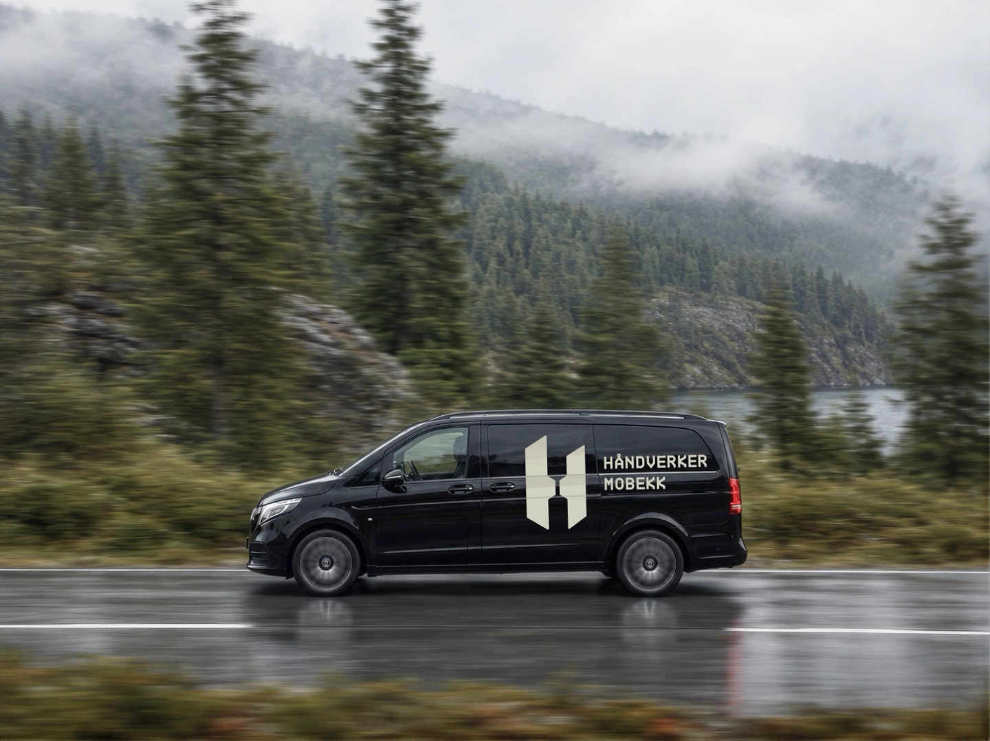 Svart varebil med Håndverker Mobekks logo og navn på siden, i fart på en våt vei med skogkledde fjell og tåke i bakgrunnen