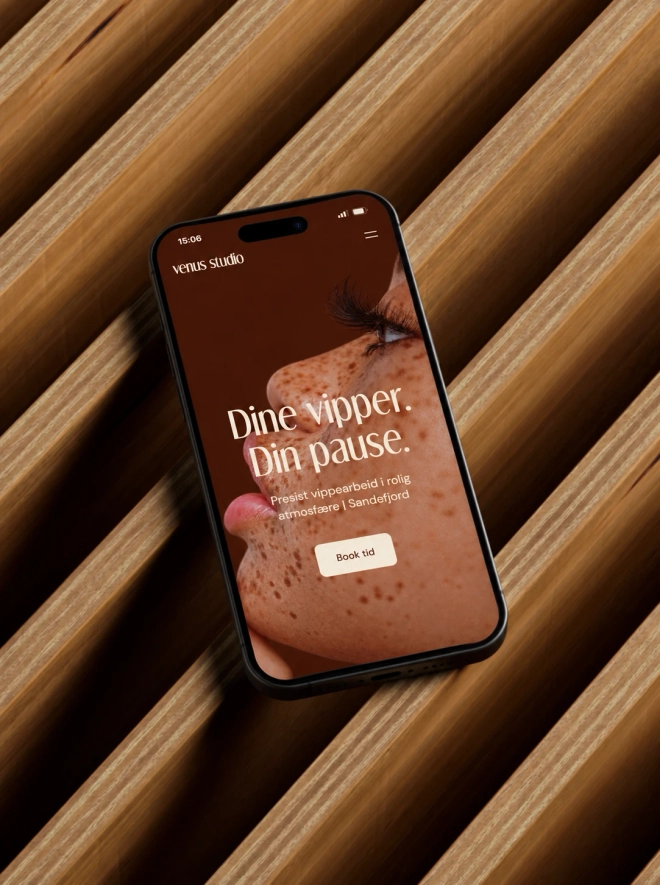 En mobiltelefon som viser Venus Studio sin nettside med overskriften «Dine vipper. Din pause.» og en knapp for å bestille time, liggende på riflede trepaneler